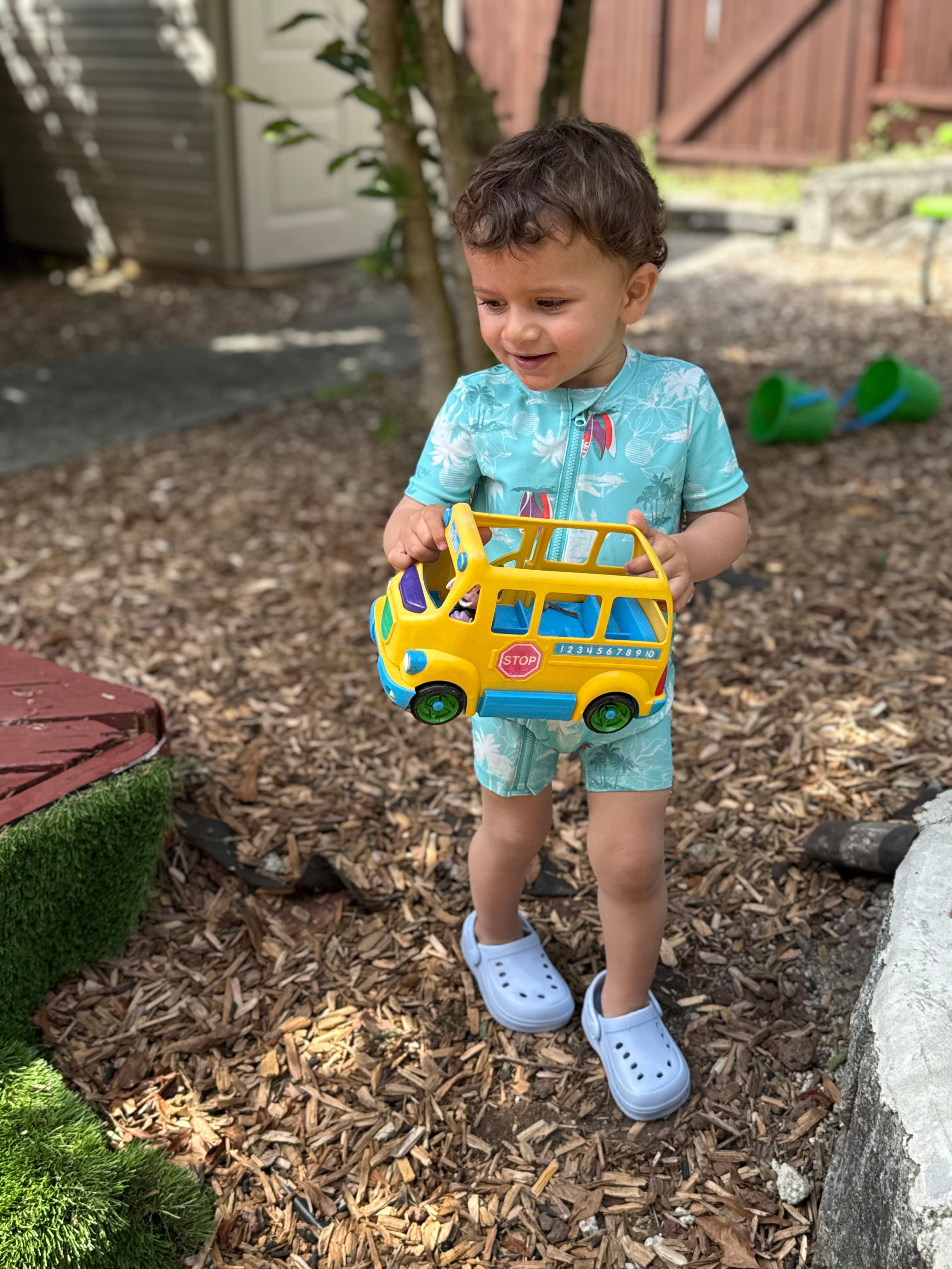 Toddler smiling outside while holding a yellow toy bus